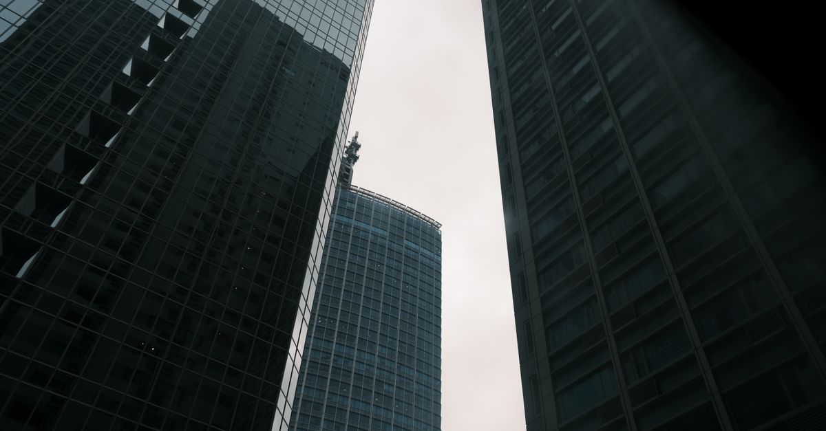 A wide-angle view of a modern city financial district at dusk, glass office towers reflecting soft light, people walking with smartphones, cinematic depth of field, no visible writing or sign, calm professional mood