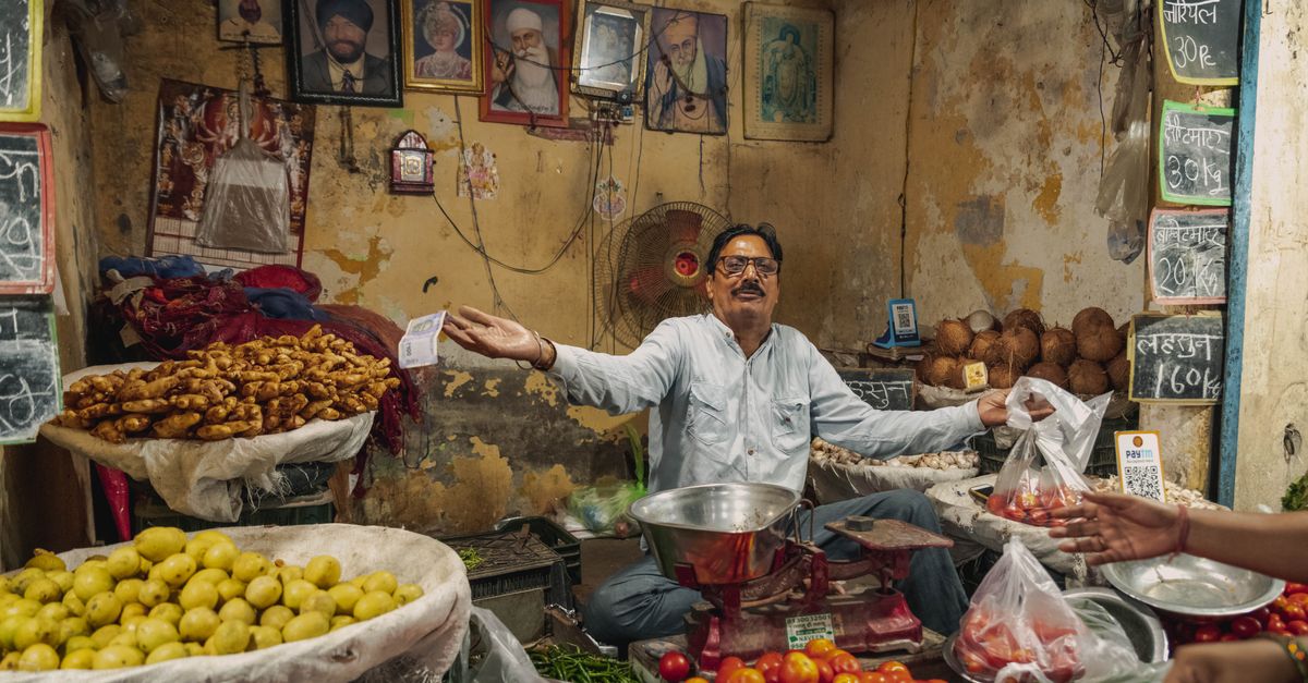 A wide-angle documentary-style view of a busy urban street in India with people using smartphones at a small market stall, warm afternoon light, no visible writing or sign, natural candid atmosphere, cinematic depth of field